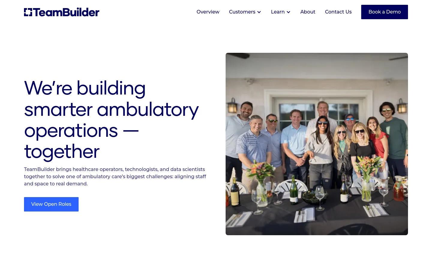 The desktop About page features a clean white background with a large headline on the left and a hero image on the right showing a group of people smiling behind a decorated outdoor table. A blue 'View Open Roles' button sits below the introductory text, and the navigation bar at the top includes links for Overview, Customers, Learn, About, Contact Us, and a 'Book a Demo' button.