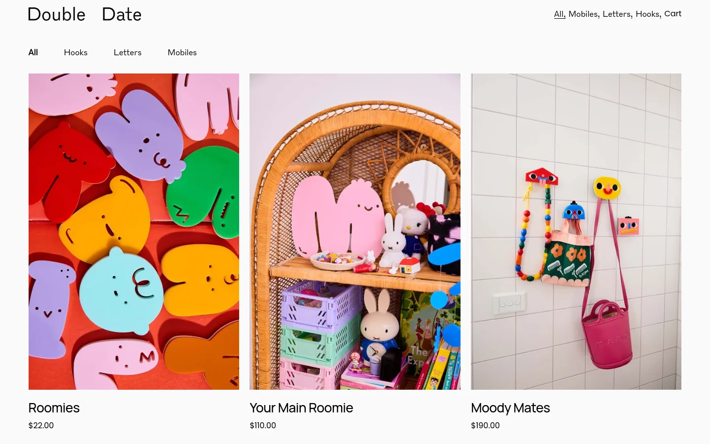 The desktop shop page for Double Date features a clean white background with a navigation menu at the top including links for All, Hooks, Letters, and Mobiles. Below the header, a grid displays colorful product images such as 'Roomies' and 'Your Main Roomie' alongside their prices. The layout is spacious with a minimalist aesthetic and high-contrast text.