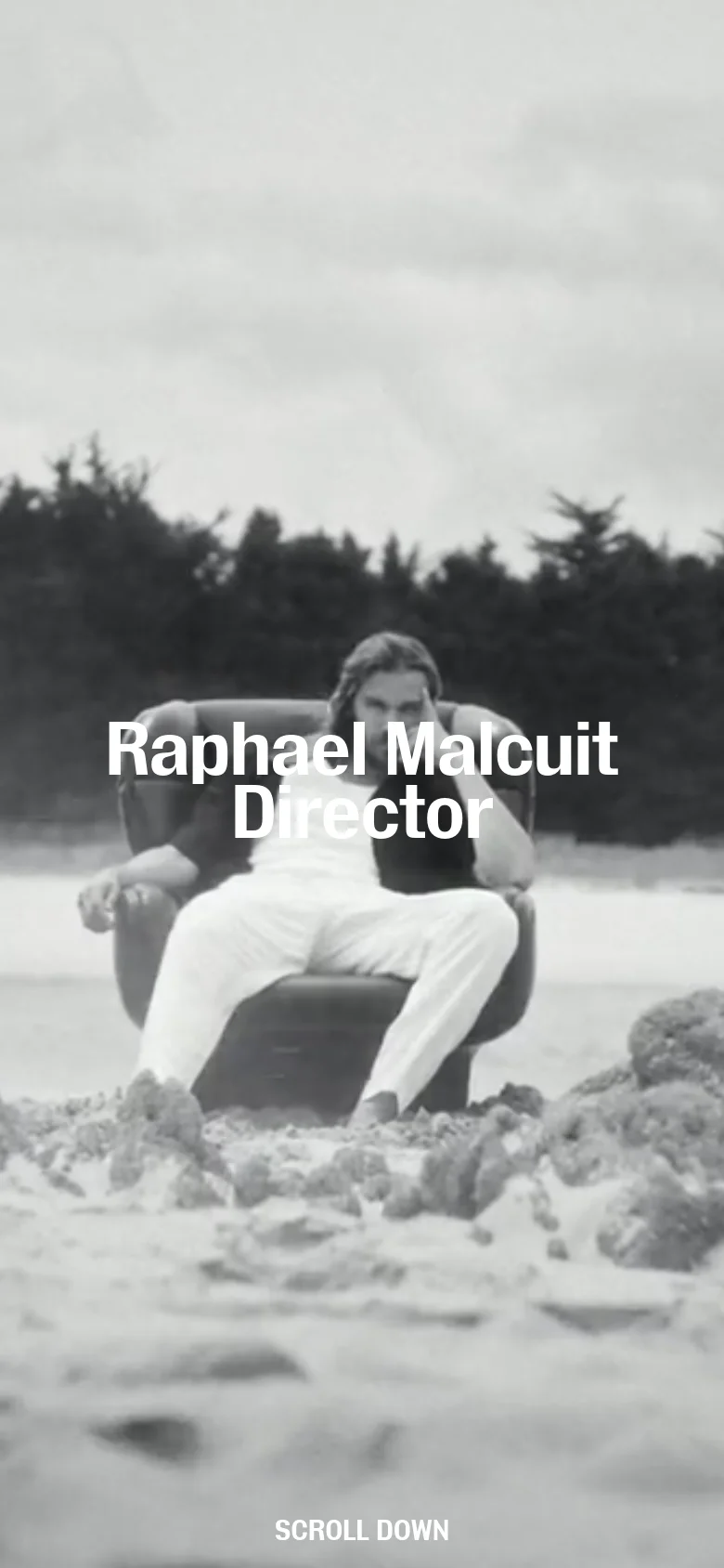 The mobile version displays a black and white version of the hero image, showing a man sitting in a chair on a beach. The centered white headline 'Raphael Malcuit Director' remains prominent, and the 'Scroll down' prompt is positioned at the bottom of the vertical frame.
