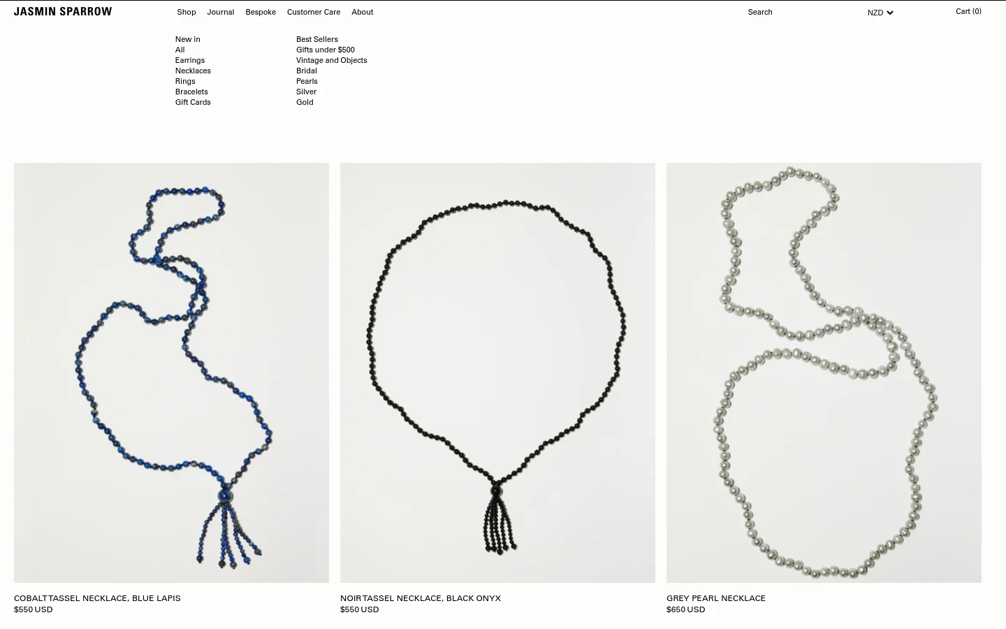 The desktop shop page features a clean, minimalist layout with a white background and black text. A navigation menu at the top includes links for jewelry categories like Earrings, Necklaces, and Rings, alongside a search bar and shopping cart. Below the menu, a grid displays high-quality product images of necklaces, such as the Cobalt Tassel Necklace and Noir Tassel Necklace, each accompanied by its name and price in USD.