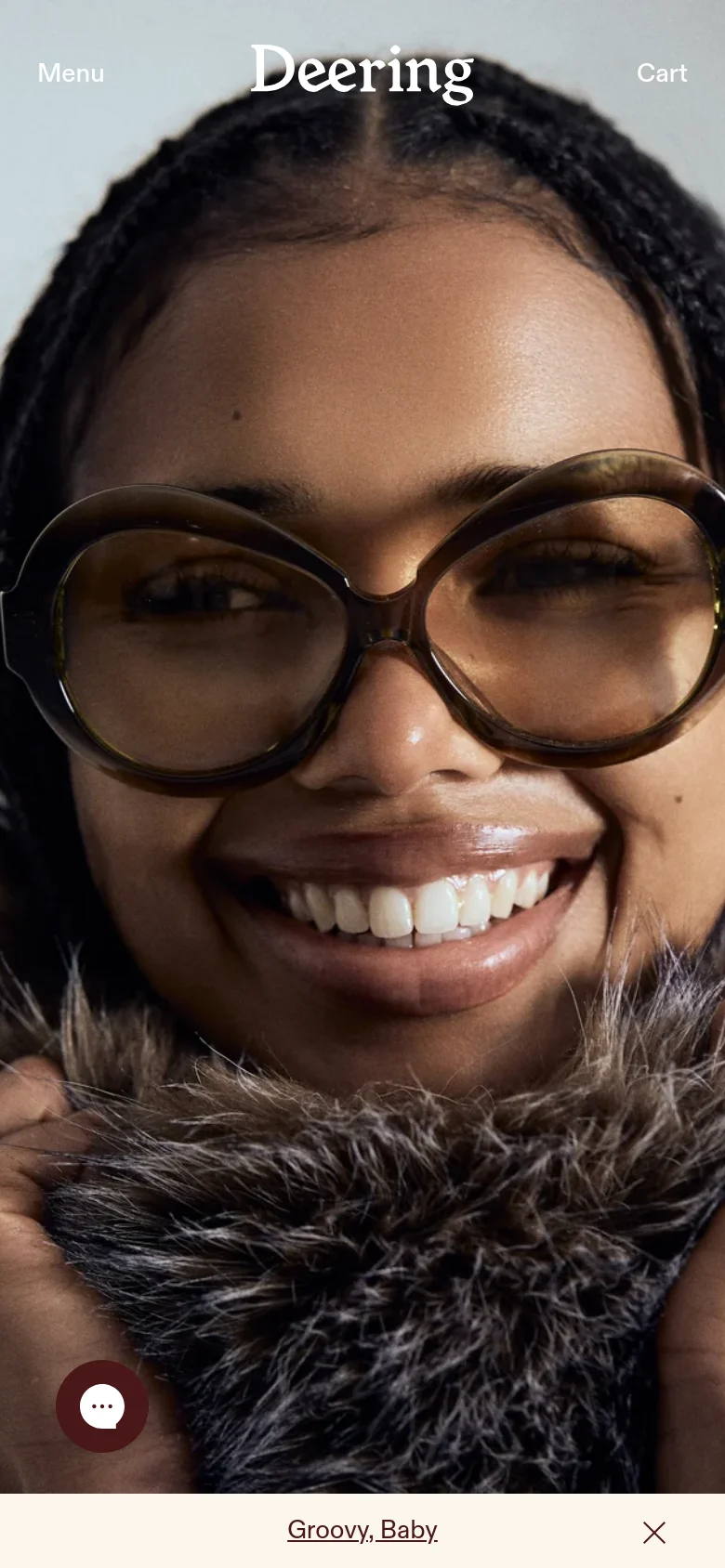 The mobile version simplifies the layout, showing the same close-up hero image of the smiling person in glasses. The top navigation is replaced by a 'Menu' button on the left and a 'Cart' button on the right, with the 'Deering' logo centered. A floating chat icon is visible in the bottom left, and a 'Groovy, Baby' banner spans the bottom of the screen.