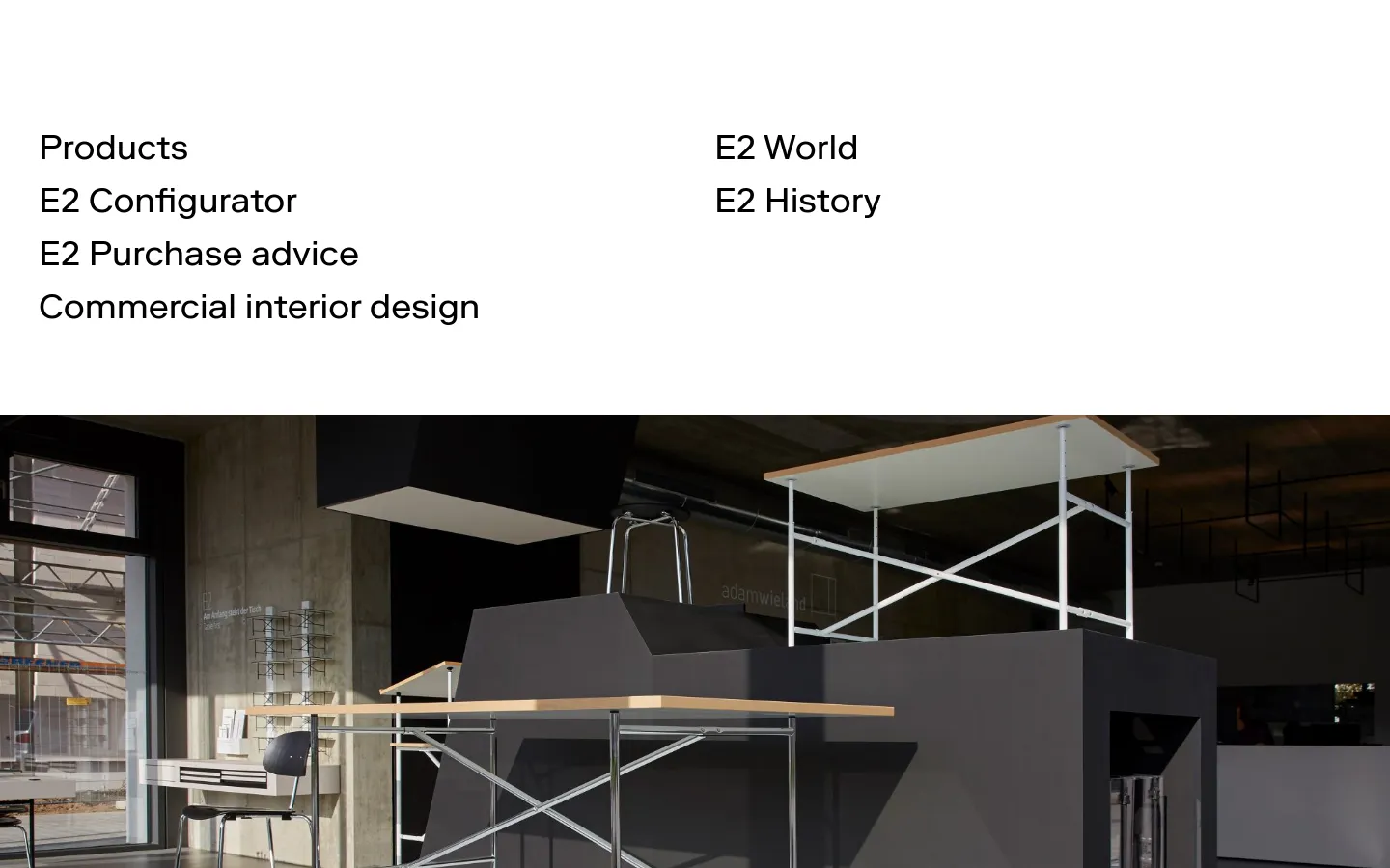 The desktop homepage features a minimalist white header with navigation links arranged in two columns. Below the navigation, a large hero image shows a modern, industrial-style interior with E2 design tables featuring metal frames and light wood tops against dark grey and concrete walls.