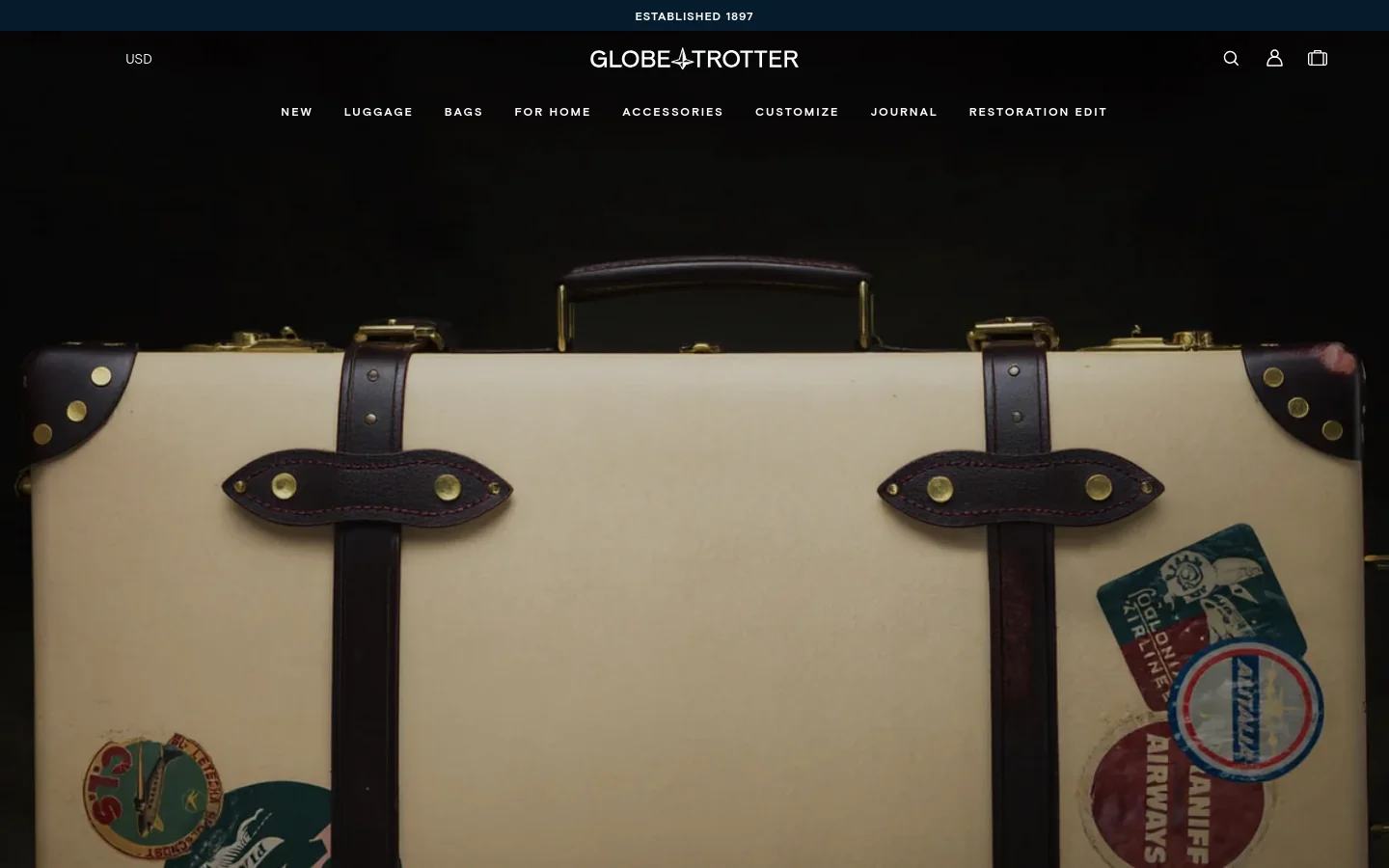 The desktop homepage features a dark, elegant header with the Globe-Trotter logo and navigation links like 'Luggage' and 'Restoration Edit'. A large hero image shows a close-up of a cream-colored vintage suitcase with leather straps and travel stickers against a dark background. The layout is spacious with a centered navigation menu and utility icons for search, account, and shopping bag.