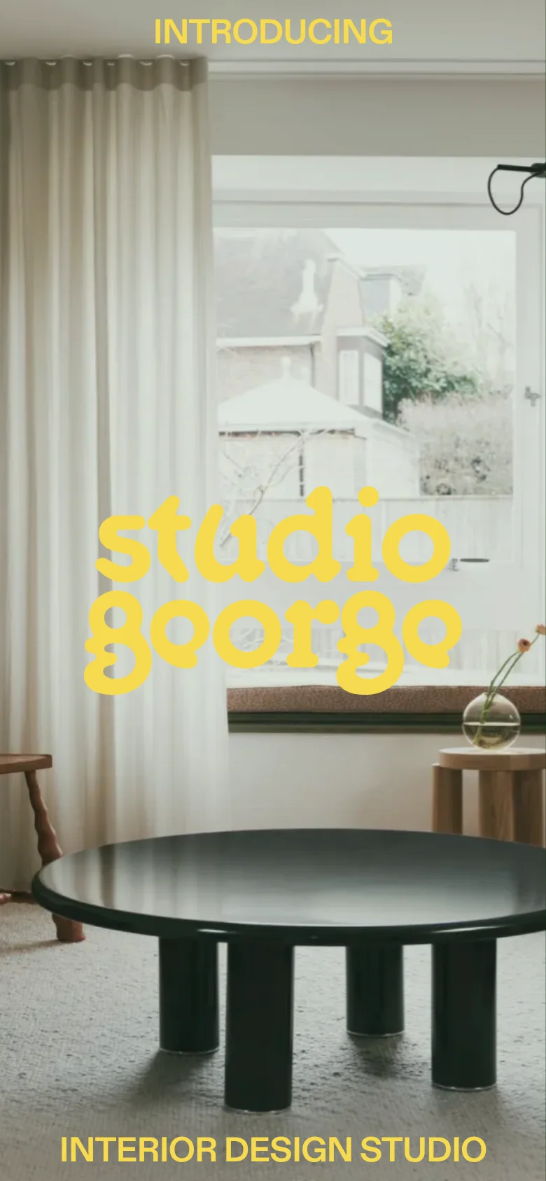 The mobile version presents a vertically oriented view of the living room hero image, focusing on a black coffee table in the foreground and a window in the background. The 'studio george' logo is centered, with the text 'INTRODUCING' at the top and 'INTERIOR DESIGN STUDIO' at the bottom.