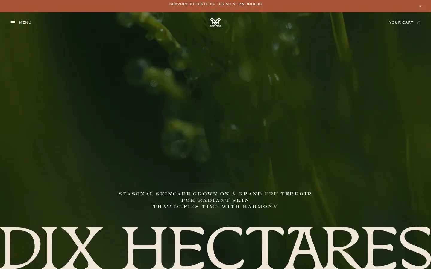 The desktop homepage features a large hero image of dew-covered green foliage with a dark, moody aesthetic. Centered white text reads 'SEASONAL SKINCARE GROWN ON A GRAND CRU TERROIR FOR RADIANT SKIN THAT DEFIES TIME WITH HARMONY' above the large brand name 'DIX HECTARES'. A top announcement bar in terracotta orange offers a promotion, and the navigation includes a menu icon on the left and a shopping cart icon on the right.