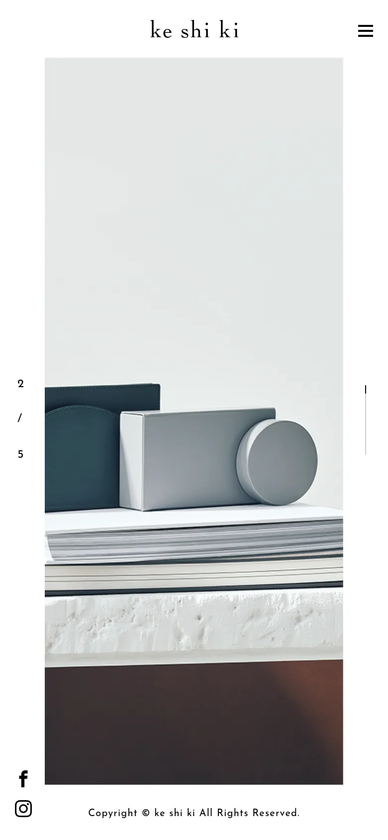 The mobile version maintains the minimalist aesthetic but shifts to a vertical layout, focusing on a large, full-width hero image of the product arrangement. The brand name is centered at the top, and the navigation is condensed into a hamburger menu icon in the top right corner. Social media icons and copyright information are placed at the bottom of the screen.