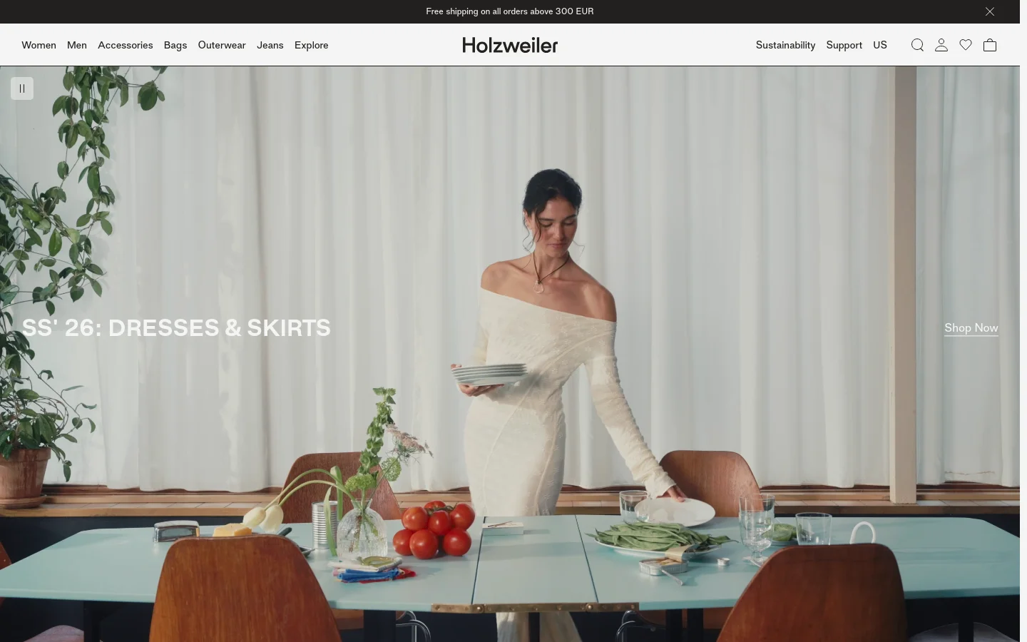 The desktop homepage features a minimalist white navigation bar with links for Women, Men, and Accessories. A large hero image shows a woman in a white off-the-shoulder dress standing behind a light blue table with tomatoes and greenery, overlaid with the headline 'SS' 26: DRESSES & SKIRTS' and a 'Shop Now' call to action.