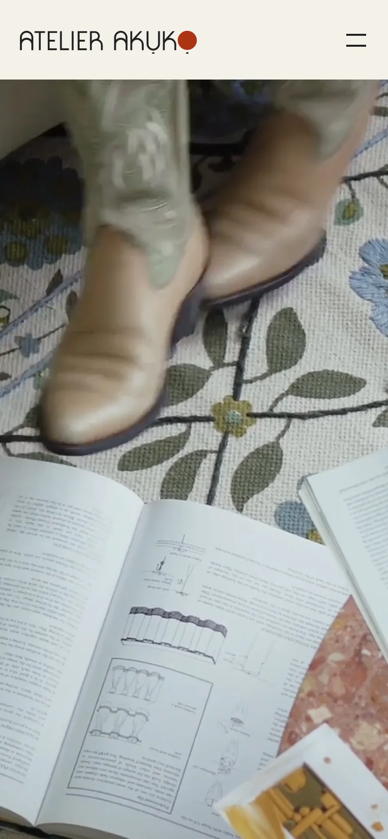 The mobile version simplifies the navigation into a hamburger menu icon next to the Atelier Akuko logo. The hero imagery transitions to a full-width, vertically oriented view of a person's legs and feet walking on a floral patterned rug next to an open book. The layout is more streamlined to accommodate the narrow screen.