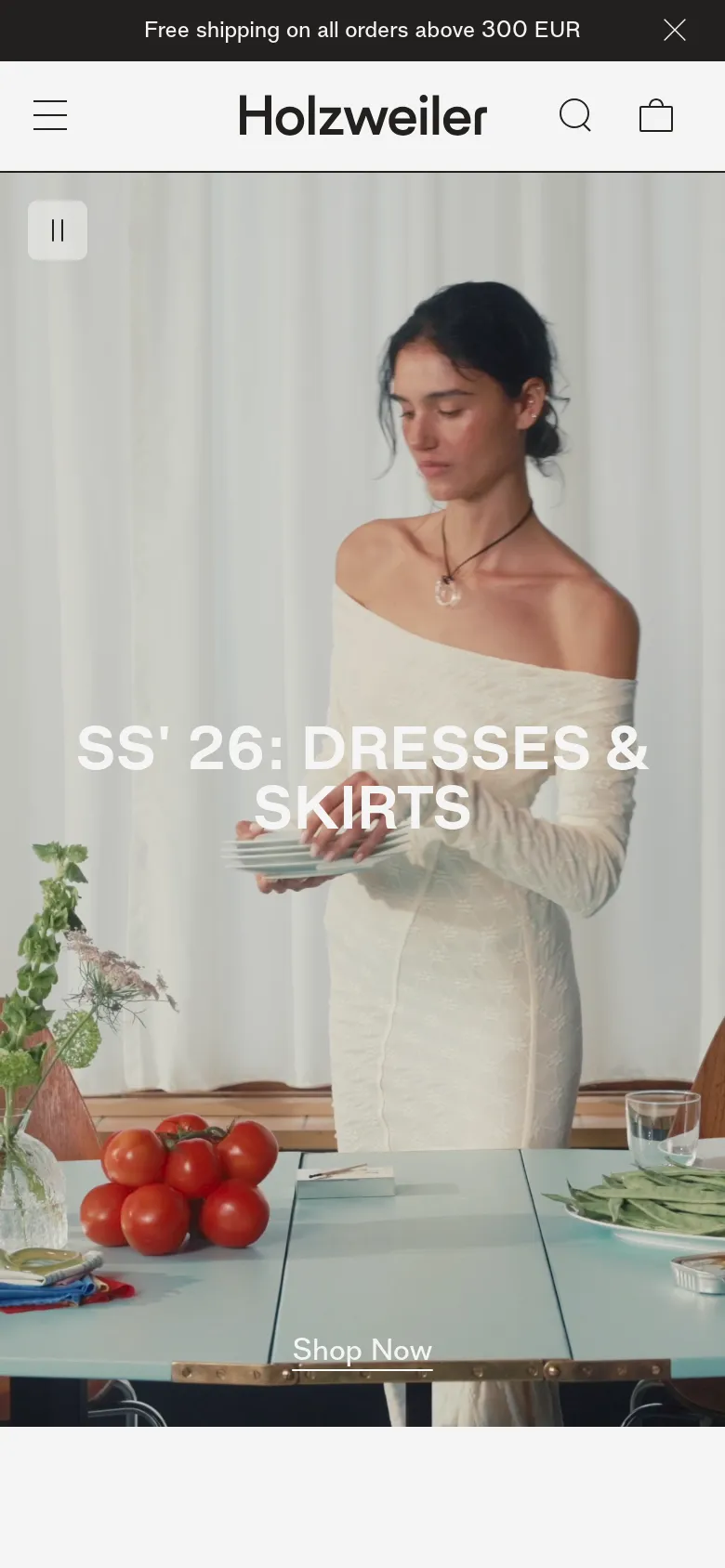 The mobile version presents a simplified layout with a hamburger menu and a centered Holzweiler logo. The hero section features the same large image of a woman in a white dress and the 'SS' 26: DRESSES & SKIRTS' headline, but the text and 'Shop Now' button are vertically stacked and centered for a smaller screen.
