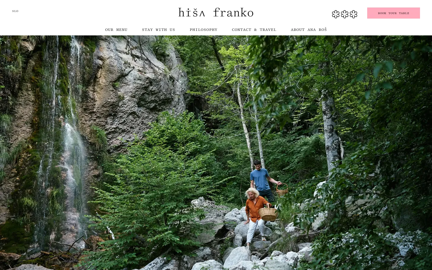 The desktop homepage features a full-width hero image of a lush, green forest landscape with a waterfall and two people walking through rocky terrain. A minimalist white navigation bar at the top contains the Hiša Franko logo, menu links like 'OUR MENU' and 'ABOUT ANA ROŠ', and a pink 'BOOK YOUR TABLE' button. The overall aesthetic is clean and nature-focused with a white and natural color palette.