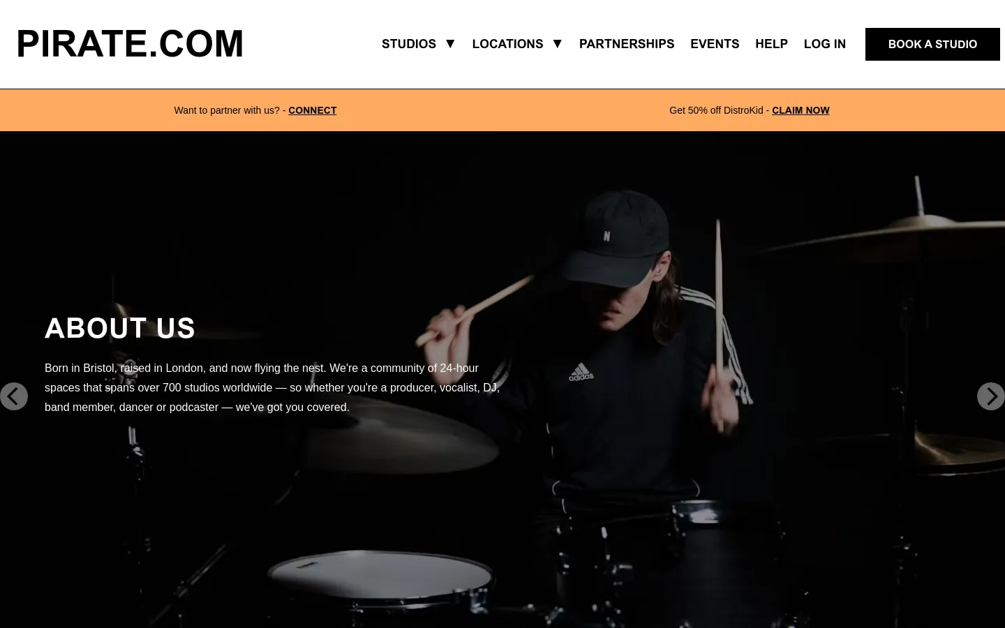 The desktop page features a large hero image of a drummer in a dark studio with the headline 'ABOUT US' and introductory text overlaid. A white navigation bar at the top includes the Pirate.com logo, menu links, and a 'BOOK A STUDIO' button, while an orange banner below offers partnership and discount links. The layout is spacious with high-contrast white text against a dark background.