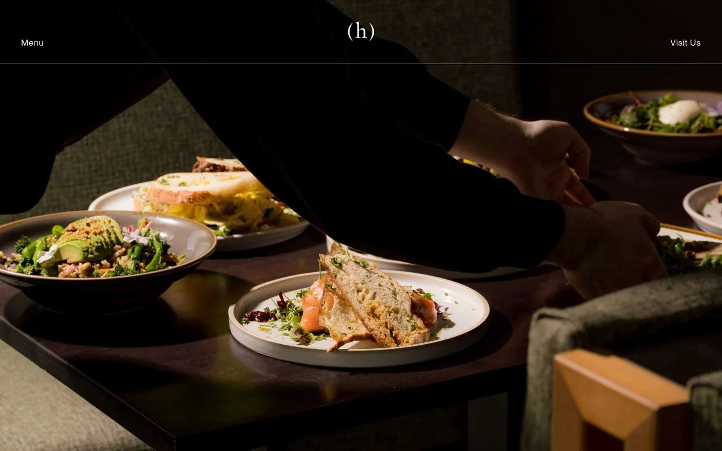 The desktop page features a dark, elegant hero image of a chef plating food on a wooden table. Below, the layout uses a clean white background with sections for 'Our menu' featuring a 'View the menu' button, and 'Reservations' with text details and a large placeholder image. The design is spacious with dark text and a minimalist navigation bar at the top containing 'Menu', a central '(h)' logo, and 'Visit Us'.