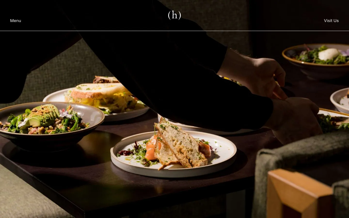 The desktop page features a dark, elegant hero image of a chef plating food on a wooden table. Below, the layout uses a clean white background with sections for 'Our menu' featuring a 'View the menu' button, and 'Reservations' with text details and a large placeholder image. The design is spacious with dark text and a minimalist navigation bar at the top containing 'Menu', a central '(h)' logo, and 'Visit Us'.