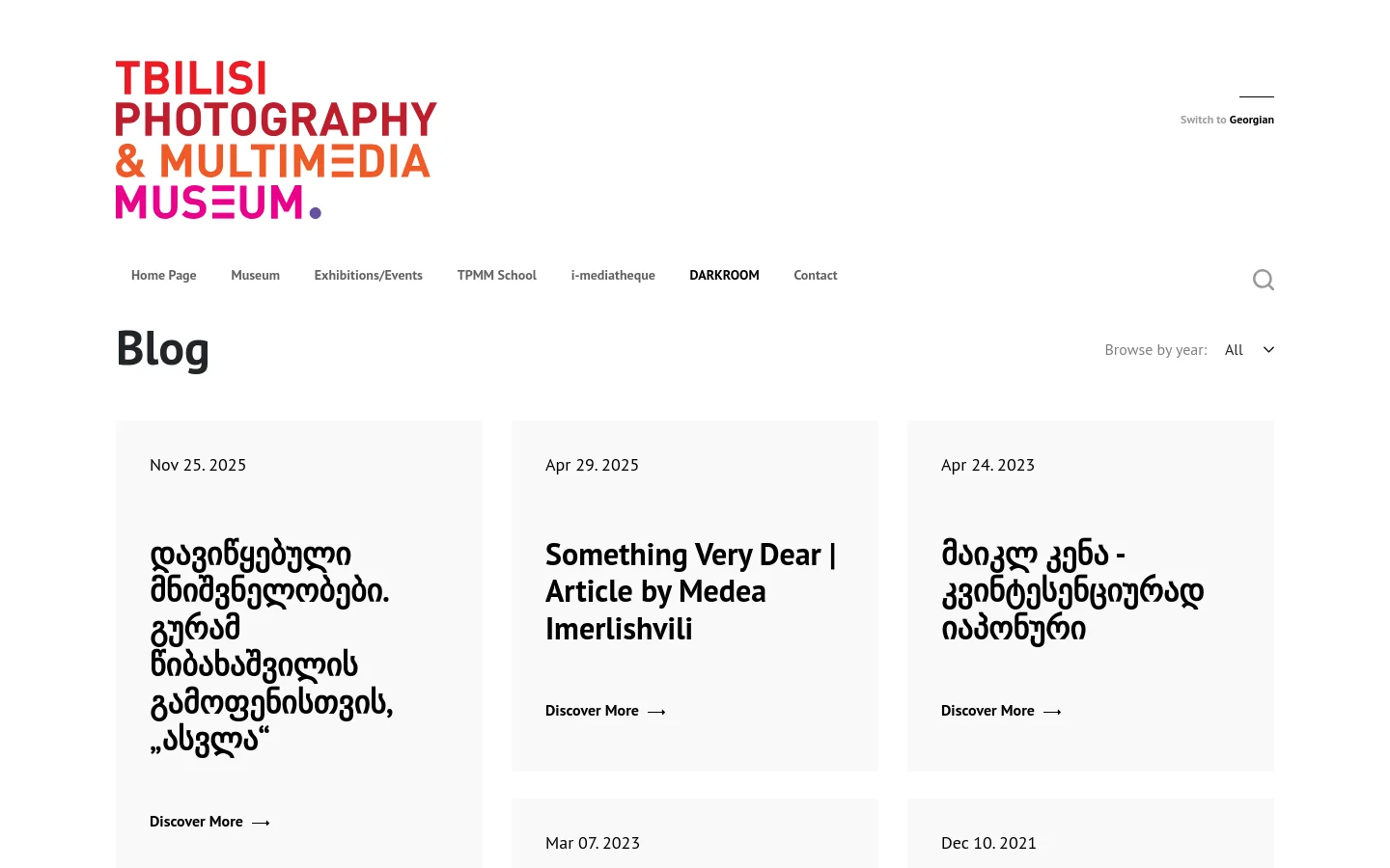 The desktop blog page for the Tbilisi Photography & Multimedia Museum features a clean white background with a large red and orange logo at the top left. A navigation menu includes links like Home Page, Museum, and DARKROOM, alongside a search icon. Below the 'Blog' heading, blog posts are arranged in a grid layout, each displaying a date, a large bold title in Georgian or English, and a 'Discover More' call to action with an arrow.