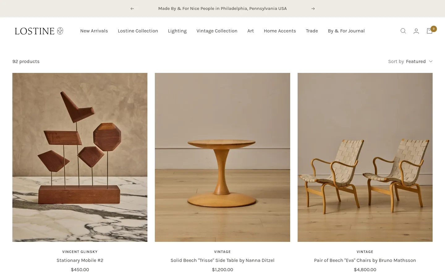 The New Arrivals page features a minimalist layout with a large lifestyle hero image at the top and a product grid below. Products like wooden mobiles, side tables, and chairs are displayed in a multi-column grid with clean typography, neutral tones, and prices listed under each item. A navigation bar at the top includes links for New Arrivals, Lighting, and Vintage Collection.