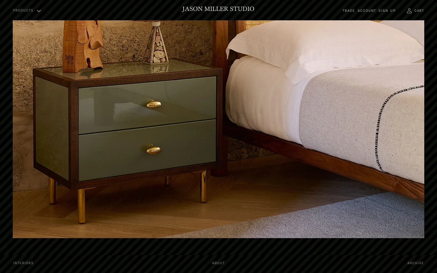 The Jason Miller Studio homepage features a minimalist design with a dark, textured border. A large hero image showcases high-end interior design, including a green two-drawer nightstand with brass legs next to a wooden bed. Navigation links for Products, Trade Account, Sign Up, and Cart are positioned at the top.