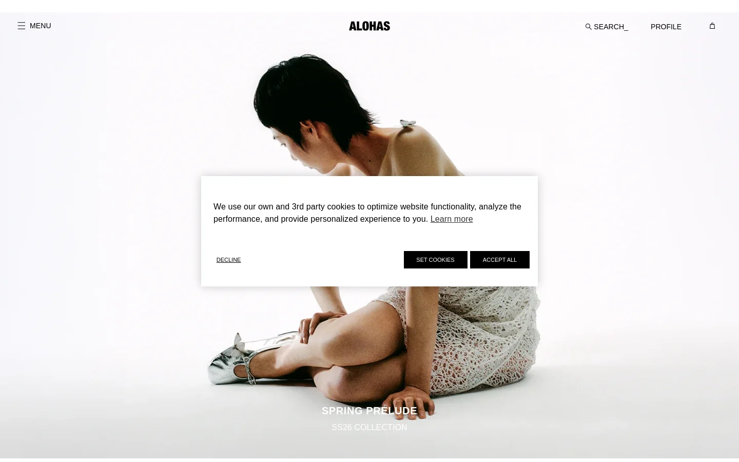 The desktop homepage features a large, minimalist hero image of a person in a white textured dress against a bright white background. A centered cookie consent overlay appears over the image, and the 'SPRING PRELUDE SS26 COLLECTION' headline is positioned at the bottom. The navigation bar is clean, containing a menu icon, the ALOHAS logo, a search function, profile link, and a shopping bag icon.