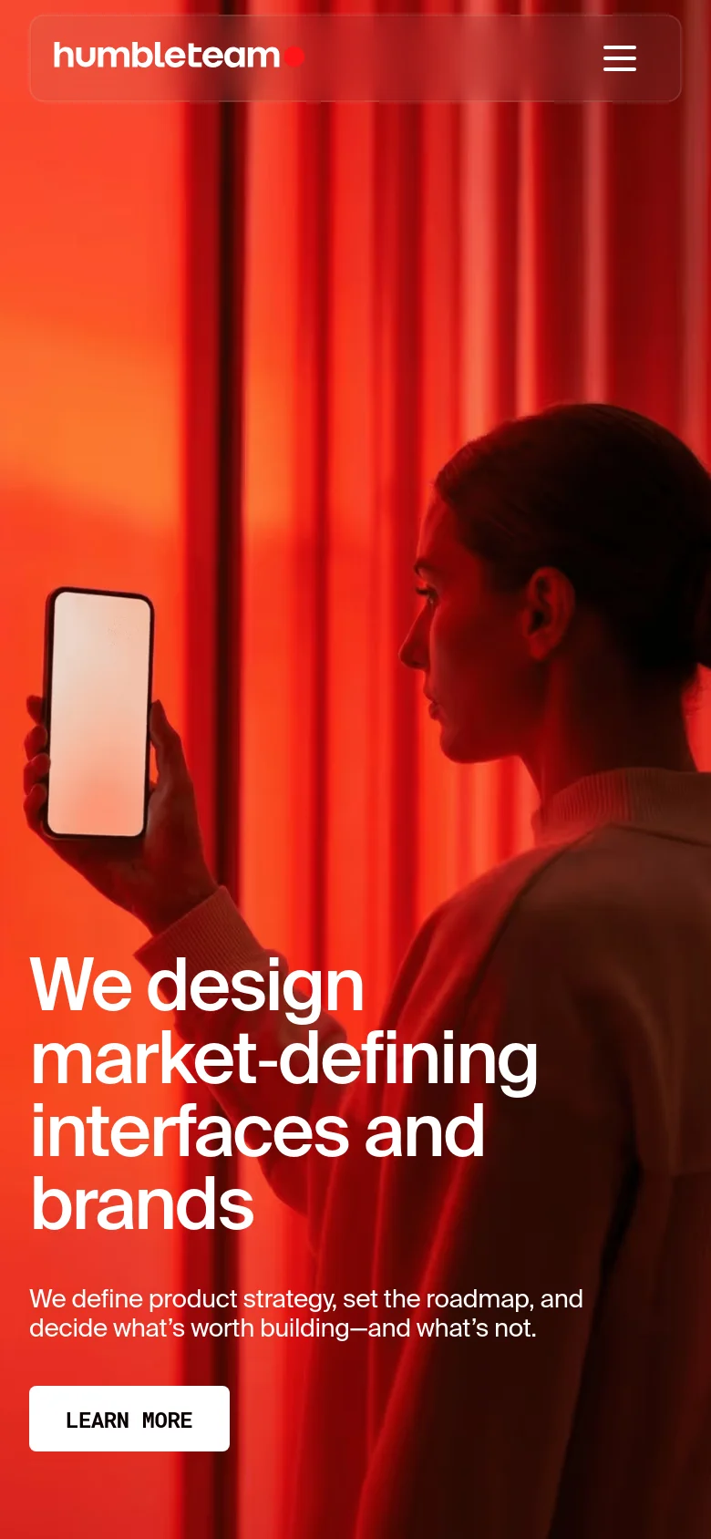 The mobile version displays a simplified layout with a red-tinted hero image of a person holding a phone. The navigation is condensed into a hamburger menu within a dark red header bar containing the Humbleteam logo. The main headline and 'Learn More' call-to-action button are stacked vertically in the center of the screen.