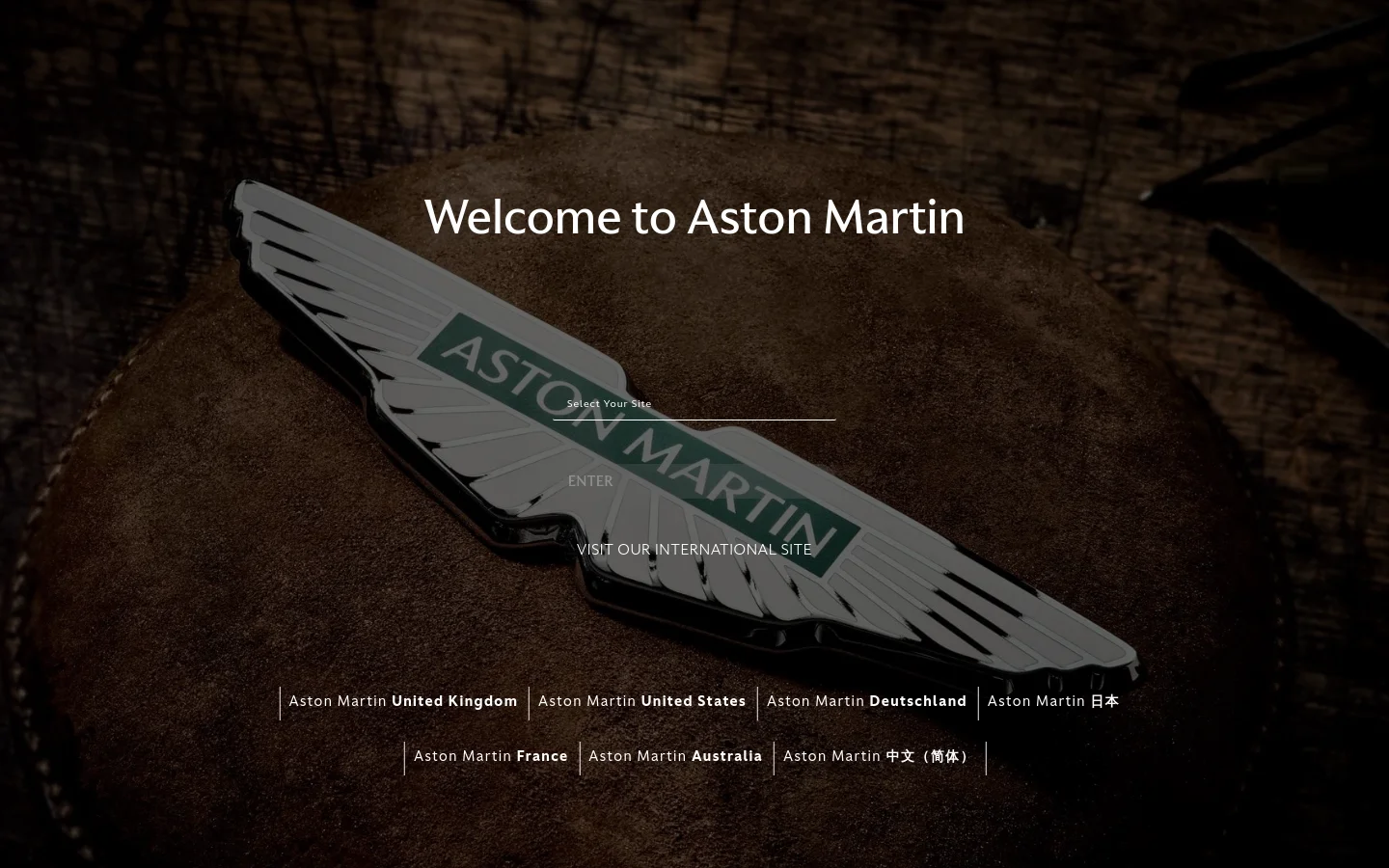 The desktop homepage features a large, close-up hero image of the silver Aston Martin winged emblem resting on a dark leather surface. Centered white text reads 'Welcome to Aston Martin' above a list of regional links for the United Kingdom, United States, Germany, Japan, France, Australia, and China.