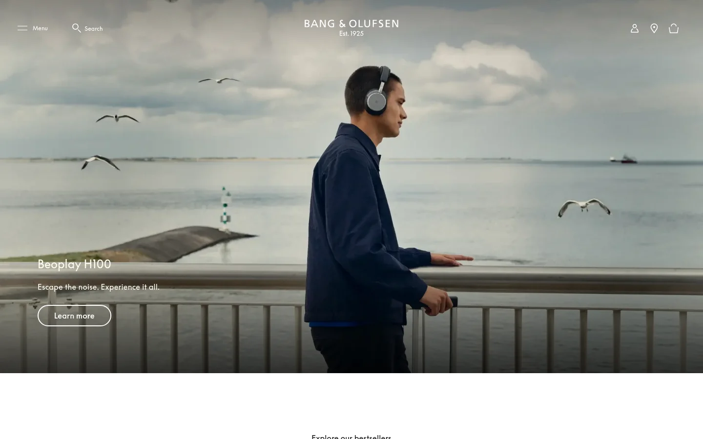 The desktop homepage features a large hero image of a man wearing Beoplay H100 headphones standing by the sea under a cloudy sky. The headline 'Beoplay H100' and a 'Learn more' call-to-action button are overlaid on the left, with a clean navigation bar at the top. Below the hero section, a grid of product bestsellers is displayed against a white background.