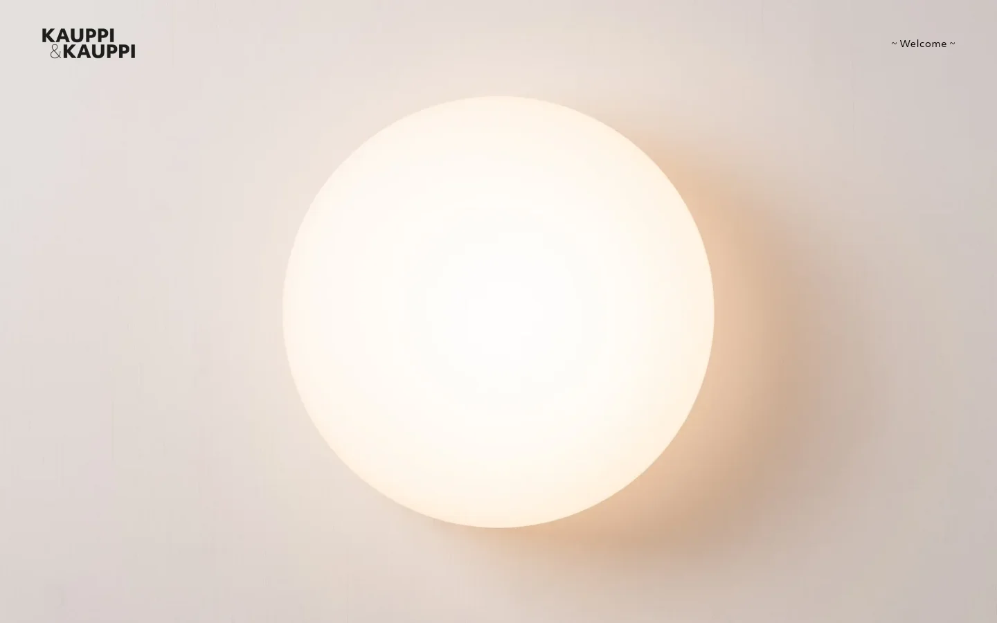 The desktop homepage features a minimalist design with a large, soft-focus hero image of a glowing white spherical lamp against a light neutral wall. The 'KAUPPI & KAUPPI' logo is positioned in the top left corner, while a '~ Welcome ~' navigation link sits in the top right. The color palette is monochromatic and warm, consisting of off-whites and soft creams.