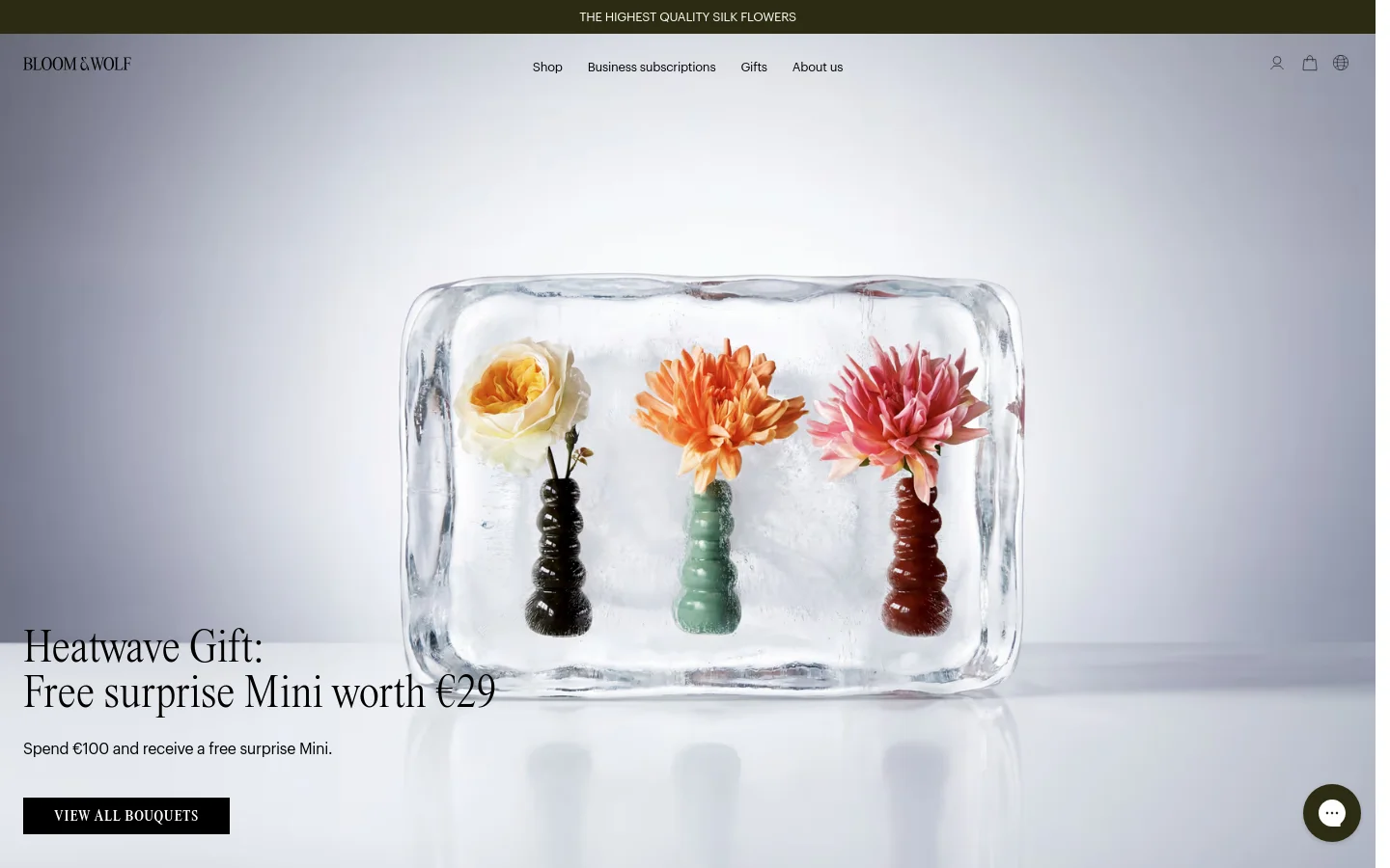 The Bloom & Wolf homepage features a large hero image of three colorful silk flowers encased in a clear ice block against a light grey background. A prominent headline announces a 'Heatwave Gift' with a call to action button labeled 'VIEW ALL BOUQUETS'. The navigation menu is clean and minimalist, set against a white header with a dark olive green promotional banner at the top.