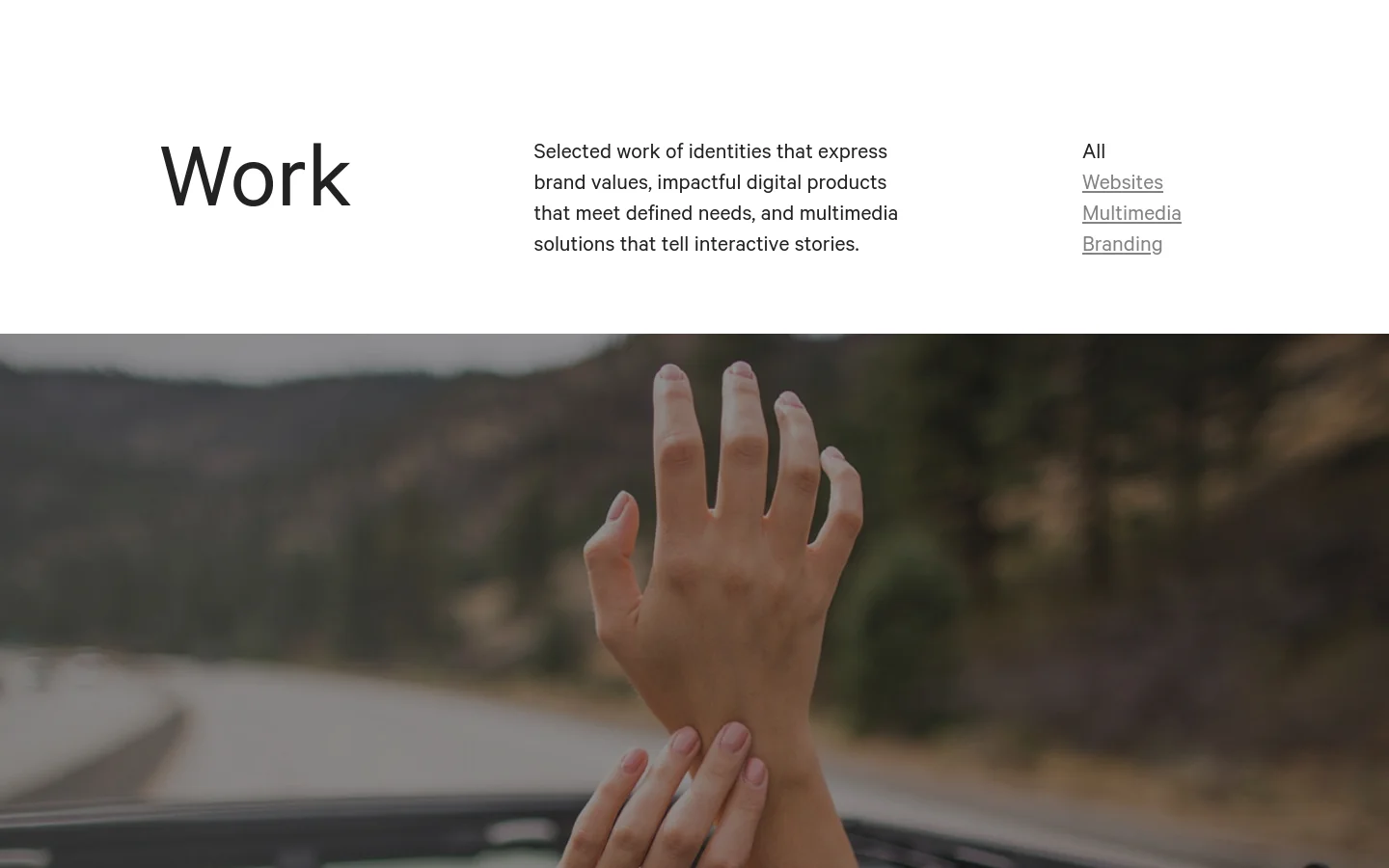 The 'Work' portfolio page features a clean, minimalist white layout with a large 'Work' headline and a brief description. A navigation menu allows filtering by All, Websites, Multimedia, or Branding, positioned to the right of the text. Below the header, a large hero image shows hands resting on a car dashboard against a blurred landscape background.