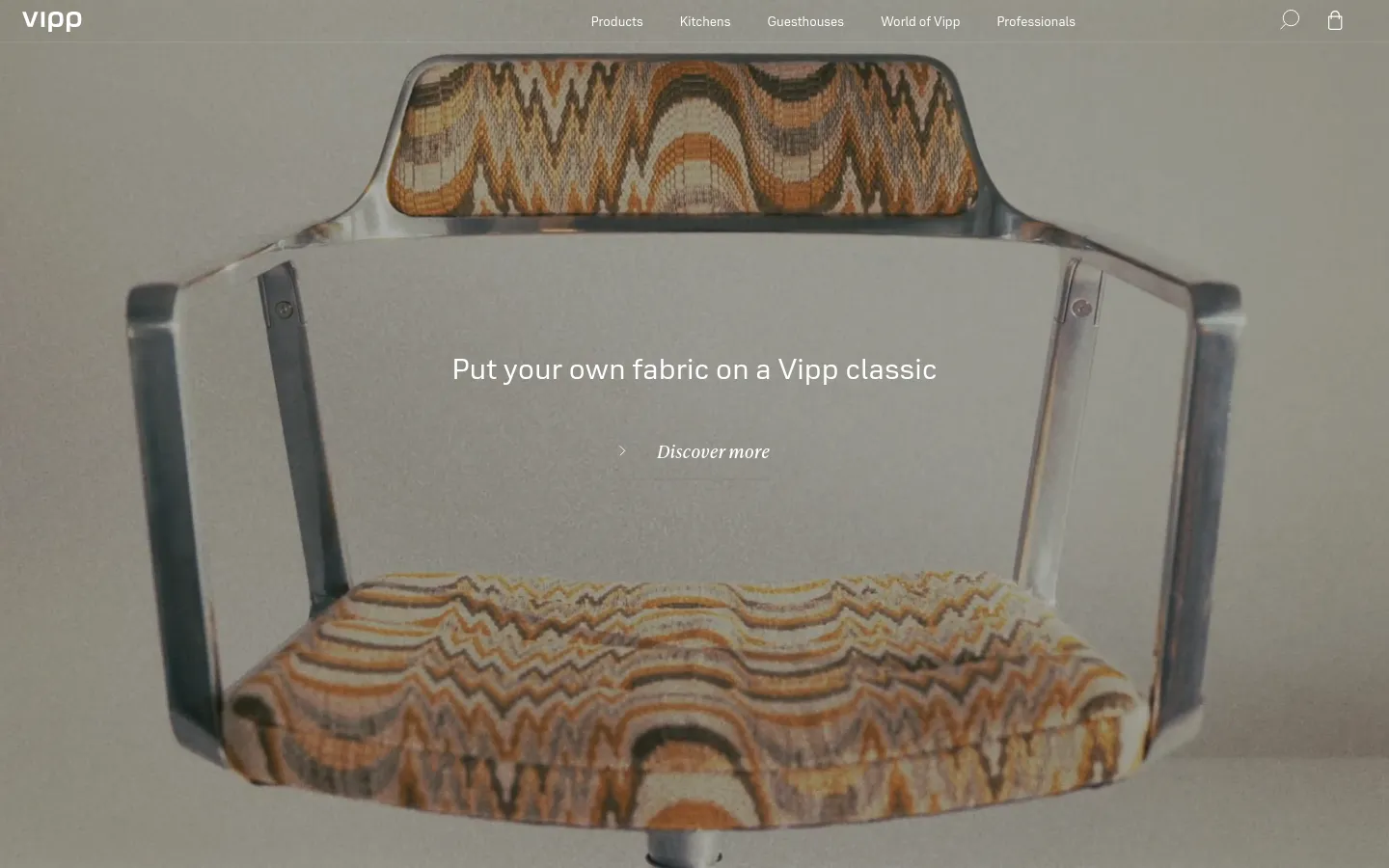 The Vipp homepage features a minimalist design with a large hero image of a modern chair with patterned fabric. The headline reads 'Put your own fabric on a Vipp classic' with a 'Discover more' call to action link. The navigation menu at the top includes links for Products, Kitchens, Guesthouses, World of Vipp, and Professionals, set against a neutral, muted background.