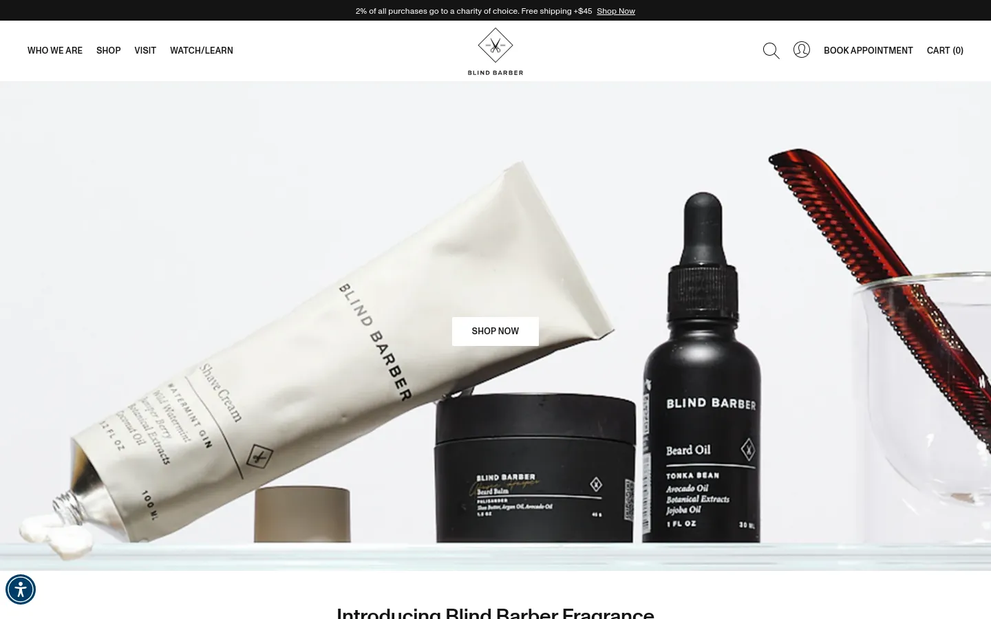 The desktop homepage features a clean, minimalist white layout with a large hero image showcasing various Blind Barber grooming products like shave cream and beard oil. A centered 'SHOP NOW' button sits over the imagery, and the navigation menu includes links for 'Who We Are', 'Shop', 'Visit', and 'Watch/Learn' alongside a search icon and account login. Below the hero section, a headline introduces the new fragrance collection.