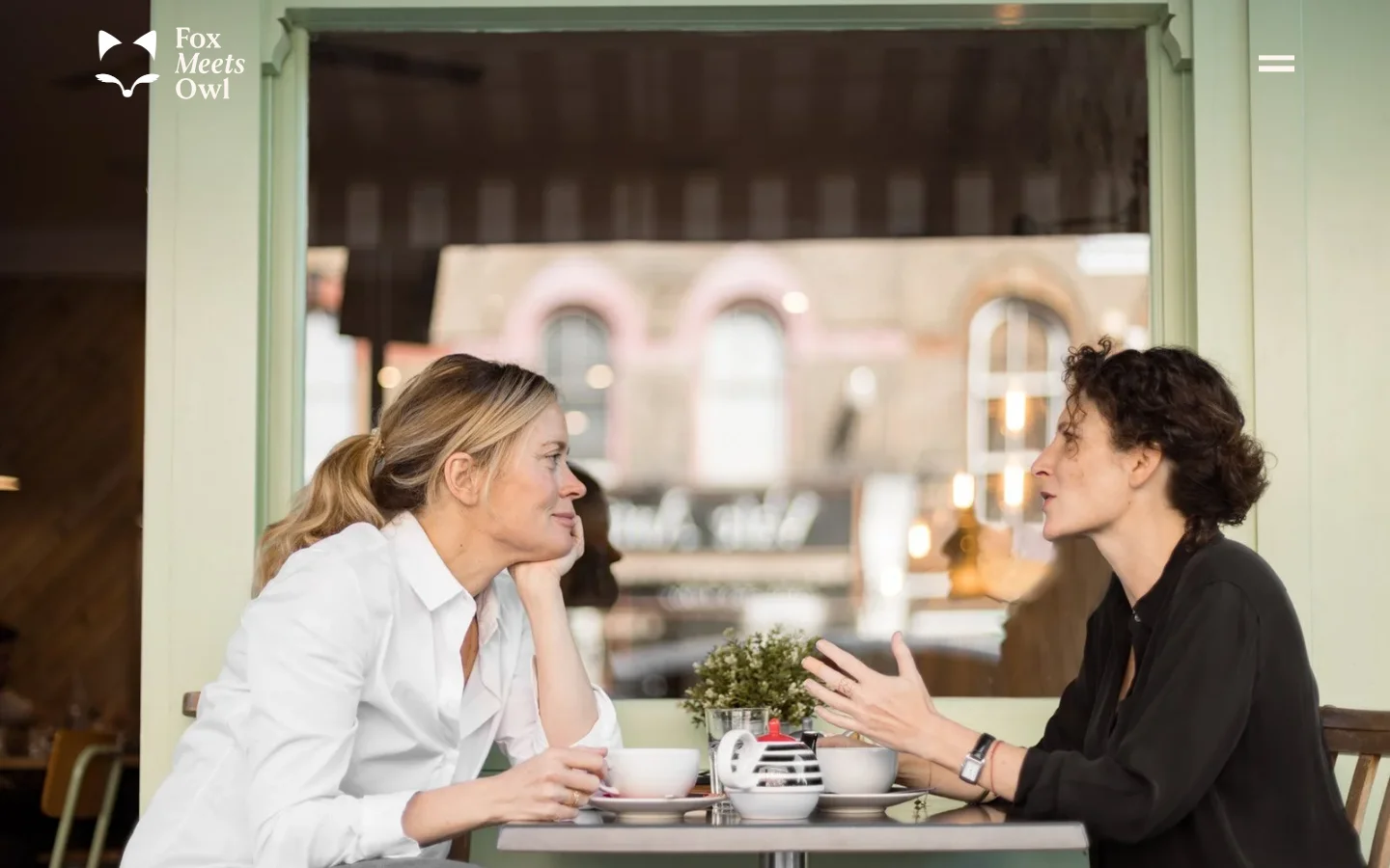 The desktop page features a large hero image of two women sitting at a cafe table engaged in conversation, set against a light green window frame. Below the image, a clean white section contains a large serif headline about leadership, followed by descriptive text and three service categories: One-on-one, Top teams, and Supervision, each accompanied by a small black icon.
