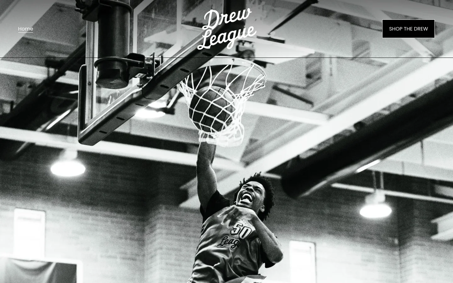The desktop homepage features a large black and white hero image of a basketball player mid-air performing a dunk. The layout includes the 'Drew League' logo in white script at the top center, a 'Home' link on the left, and a 'SHOP THE DREW' button on the right, all overlaid on the high-contrast action shot.