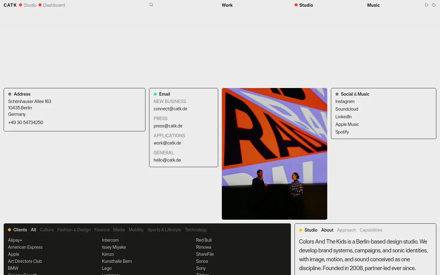 The desktop page features a minimalist, light gray layout with content organized into clean, bordered white modules. Sections include contact details, email categories, and social links, alongside a large, vibrant abstract photograph with orange and blue tones. A dark footer contains a comprehensive list of clients and studio information.