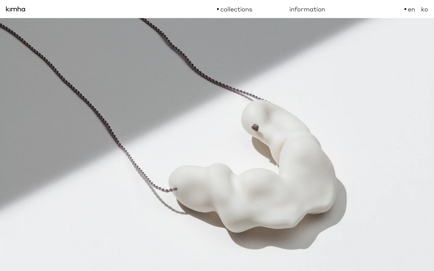 The desktop page features a minimalist white background with a large hero image of a white, organic-shaped sculptural necklace on a thin chain. A simple navigation bar at the top includes the brand name 'kimha', links for 'collections' and 'information', and language selectors. Below the hero image, a list of collection names like 'Audiable' and 'Some' serves as text-based navigation links.