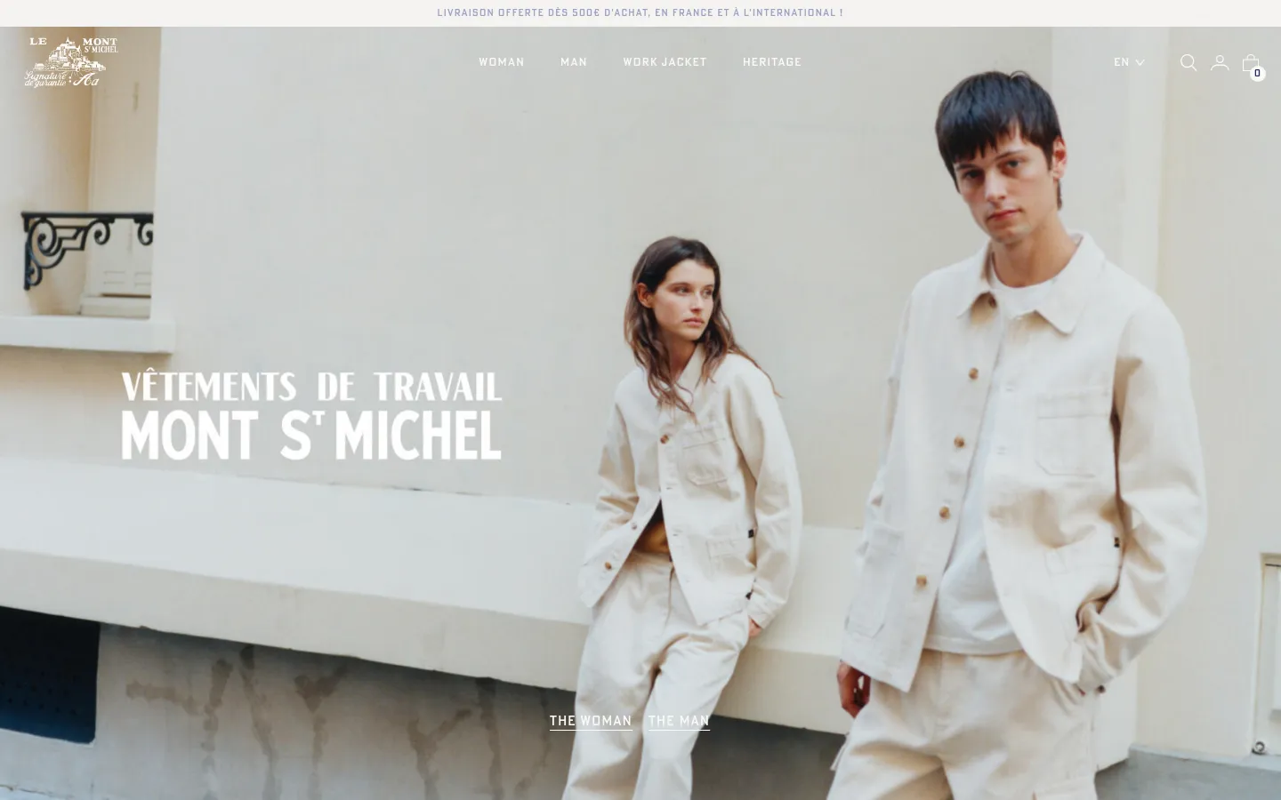 The desktop homepage features a large hero image of a man and woman in cream-colored workwear against a light wall, with the text 'VÊTEMENTS DE TRAVAIL MONT ST MICHEL' overlaid. A clean navigation menu at the top includes links for Woman, Man, Work Jacket, and Heritage, alongside a brand logo and utility icons. The layout is spacious with a minimalist aesthetic and a neutral color palette.