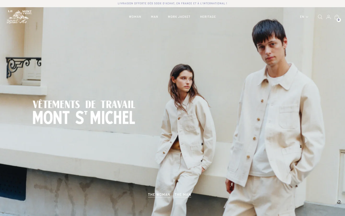 The desktop homepage features a large hero image of a man and woman in cream-colored workwear against a light wall, with the text 'VÊTEMENTS DE TRAVAIL MONT ST MICHEL' overlaid. A clean navigation menu at the top includes links for Woman, Man, Work Jacket, and Heritage, alongside a brand logo and utility icons. The layout is spacious with a minimalist aesthetic and a neutral color palette.