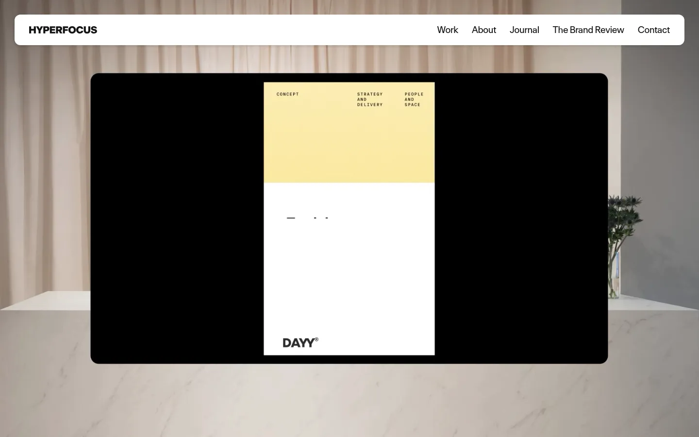 The desktop homepage features a minimalist white navigation bar at the top with the Hyperfocus logo and links for Work, About, Journal, The Brand Review, and Contact. The hero section displays a large, centered image of a design project on a black screen, set against a soft-focus background of a marble surface and draped fabric. The overall aesthetic is clean, modern, and high-end with a neutral color palette.