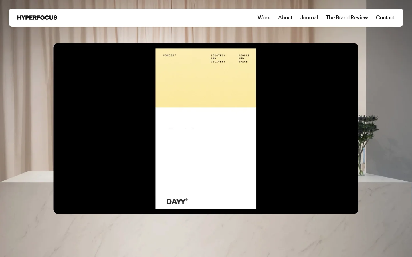 The desktop homepage features a minimalist white navigation bar at the top with the Hyperfocus logo and links for Work, About, Journal, The Brand Review, and Contact. The hero section displays a large, centered image of a design project on a black screen, set against a soft-focus background of a marble surface and draped fabric. The overall aesthetic is clean, modern, and high-end with a neutral color palette.