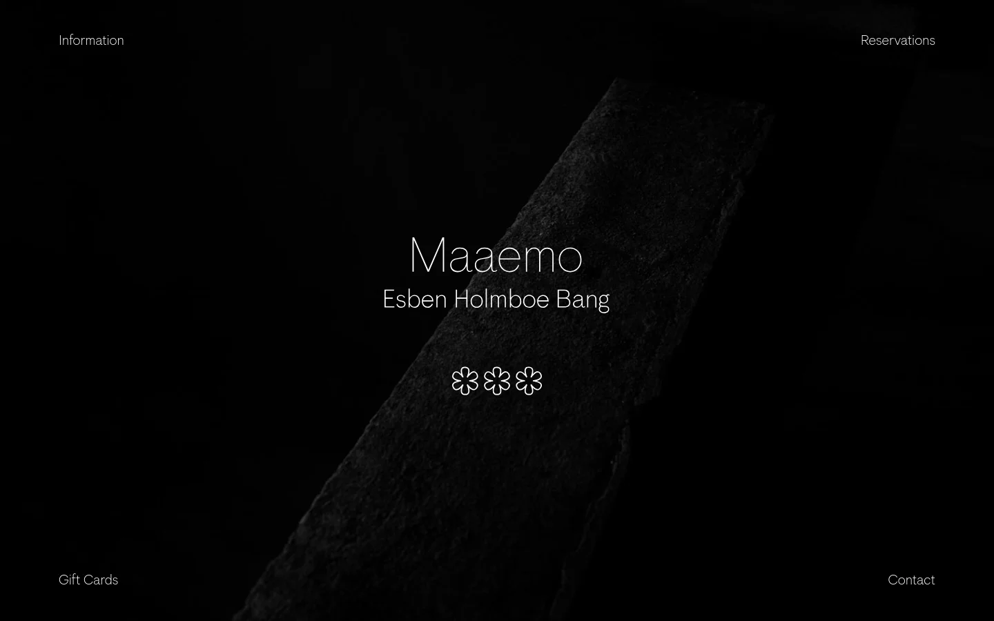 The desktop homepage features a dark, minimalist aesthetic with a large, textured dark stone element diagonally crossing the frame. Centered white text displays 'Maaemo' followed by 'Esben Holmboe Bang' and three flower icons. Navigation links for 'Information' and 'Reservations' are at the top, while 'Gift Cards' and 'Contact' are at the bottom.
