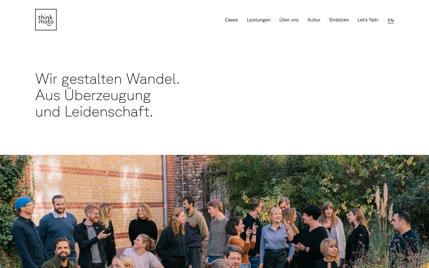 The desktop page features a minimalist white header with the think moto logo and navigation links. A large headline reads 'Wir gestalten Wandel. Aus Überzeugung und Leidenschaft.' above a wide, cinematic photograph of a diverse group of people talking in an outdoor garden setting with brick walls and greenery. The color palette is clean with high-contrast black text on a white background.