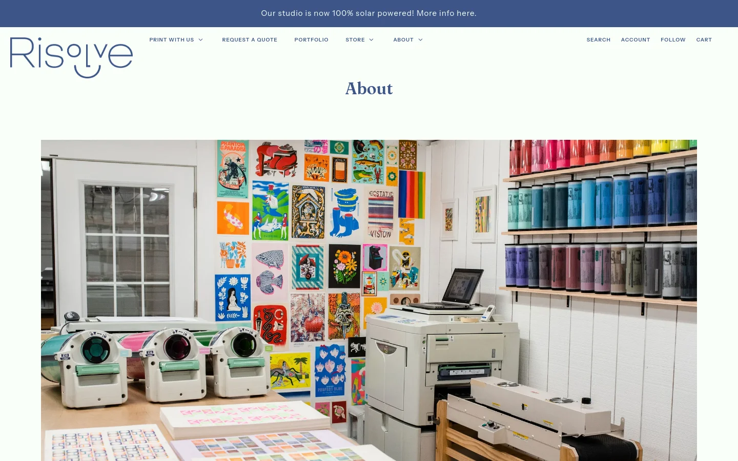 The About page features a clean white layout with a blue announcement bar at the top. A large hero image shows a bright print studio filled with Risograph machines, colorful art prints on the wall, and rows of ink canisters. Below the image, centered serif text describes the studio's history and mission.