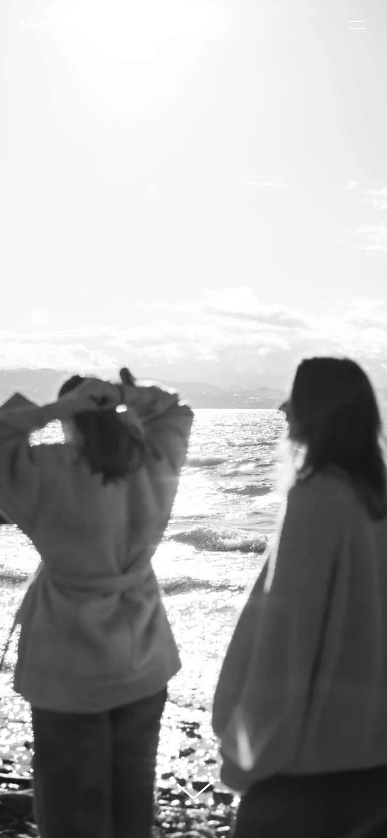 The mobile version presents a full-screen, vertical black-and-white hero image of two women by the ocean, emphasizing the serene atmosphere. The navigation is condensed into a hamburger menu icon in the top right corner, and a downward-pointing arrow at the bottom encourages scrolling through the text content.