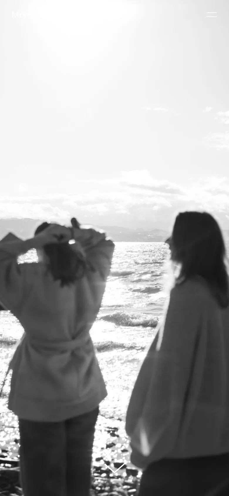 The mobile version presents a full-screen, vertical black-and-white hero image of two women by the ocean, emphasizing the serene atmosphere. The navigation is condensed into a hamburger menu icon in the top right corner, and a downward-pointing arrow at the bottom encourages scrolling through the text content.