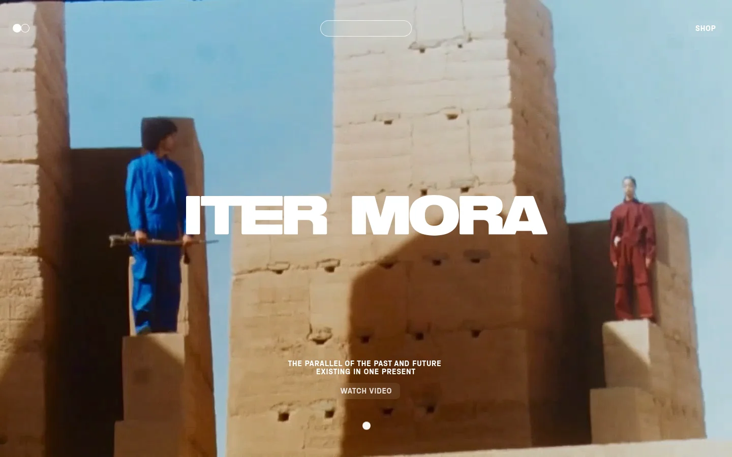 The ITER MORA homepage features a large hero image of people in colorful jumpsuits standing against ancient stone structures under a blue sky. Centered white text reads 'ITER MORA' above the tagline 'THE PARALLEL OF THE PAST AND FUTURE EXISTING IN ONE PRESENT', with a 'WATCH VIDEO' button below. Navigation includes a 'SHOP' link and a minimalist menu icon.