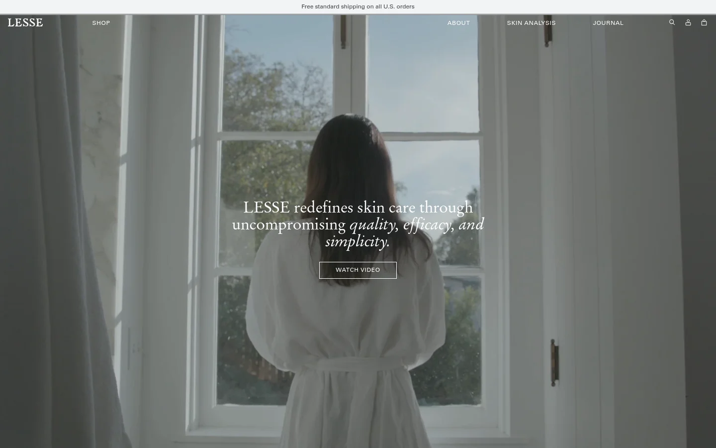 The desktop page features a minimalist design with a large hero image of a woman in a white robe looking out a window, overlaid with the headline 'LESSE redefines skin care through uncompromising quality, efficacy, and simplicity' and a 'WATCH VIDEO' button. A white navigation bar at the top includes links for Shop, About, Skin Analysis, and Journal. The color palette is neutral and airy with soft whites and natural light.