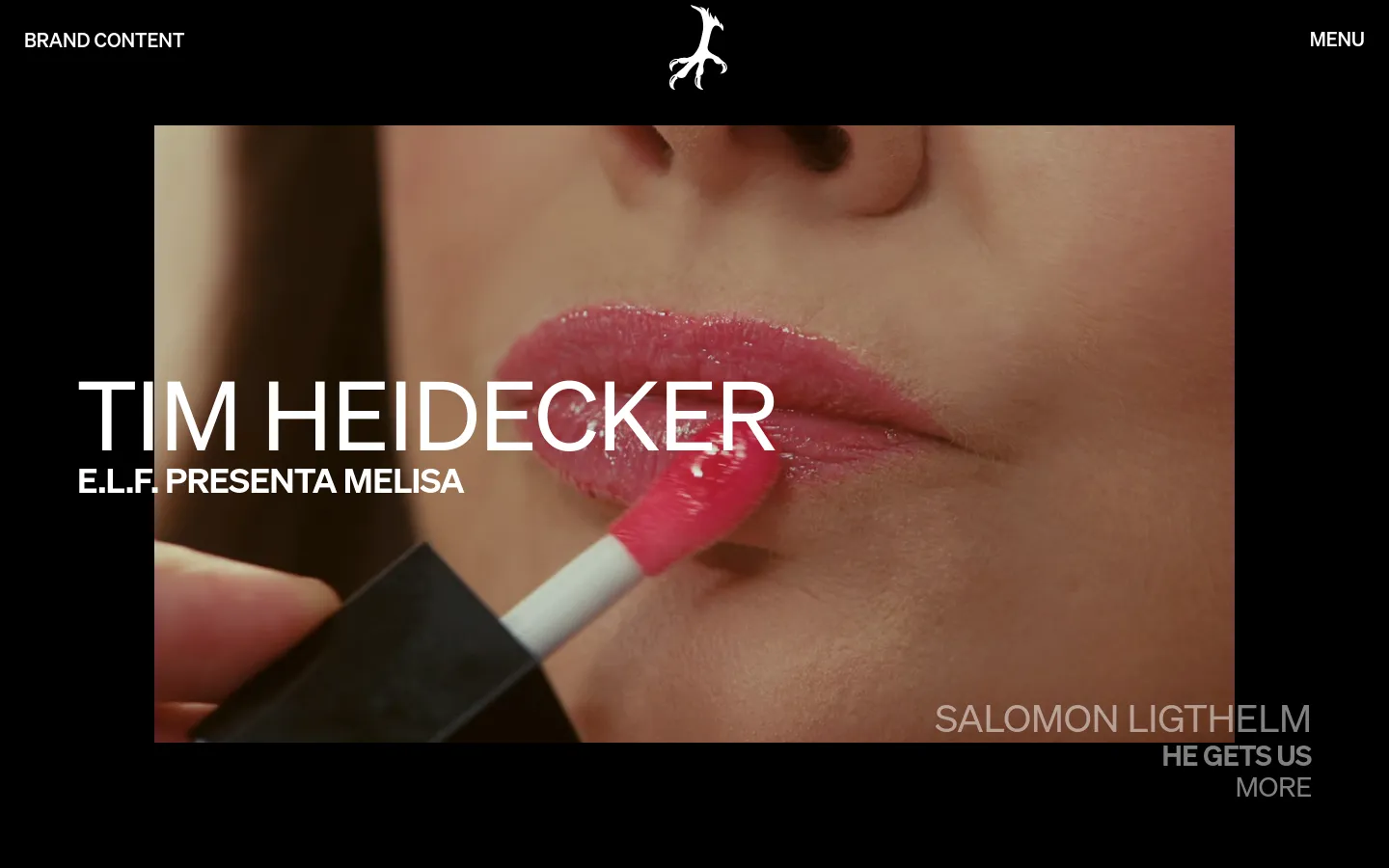 The desktop page features a dark theme with a large, cinematic hero image showing a close-up of lips being applied with pink gloss. Large white text overlays the image reading 'TIM HEIDECKER' and 'E.L.F. PRESENTA MELISA', with 'SALOMON LIGTHELM' and 'HE GETS US MORE' positioned in the bottom right. The header contains the page title 'BRAND CONTENT' on the left, a central white bird logo, and a 'MENU' button on the right.