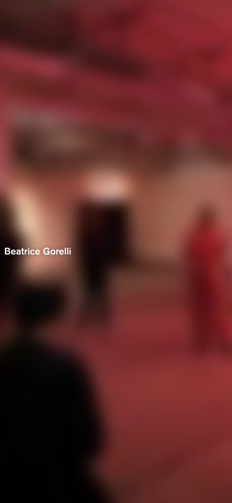 The mobile version maintains the dark aesthetic and minimalist layout, but the text 'Beatrice Gorelli' is centered more prominently. The background image is a highly blurred, warm-toned reddish abstract shape that fills the entire screen.