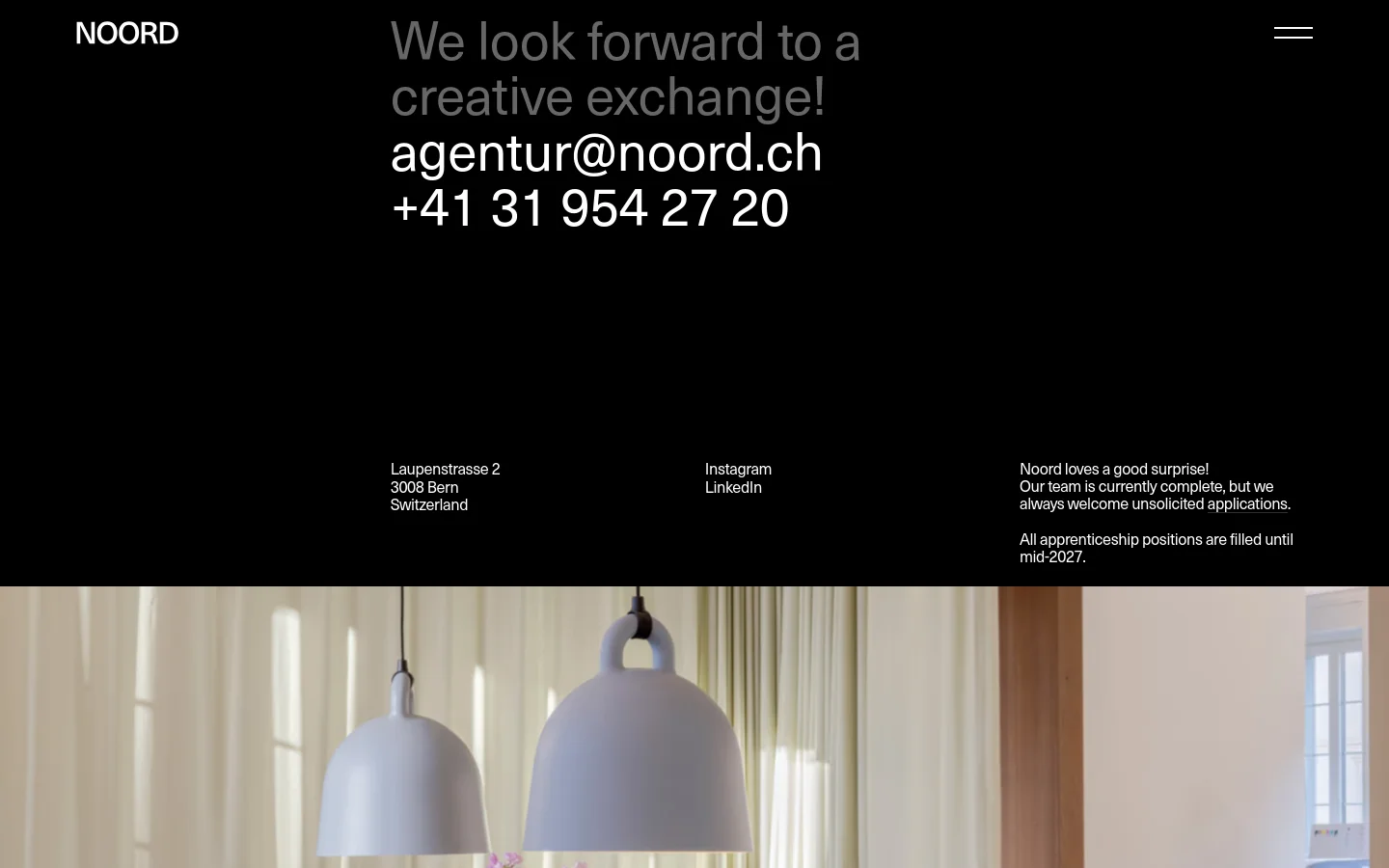 The contact page features a minimalist dark theme with white text. A large headline reads 'We look forward to a creative exchange!' above the agency's email and phone number. Below, the layout is divided into columns containing the physical address, social media links, and a note regarding job applications, all positioned above a large lifestyle photograph of an interior with pendant lights.