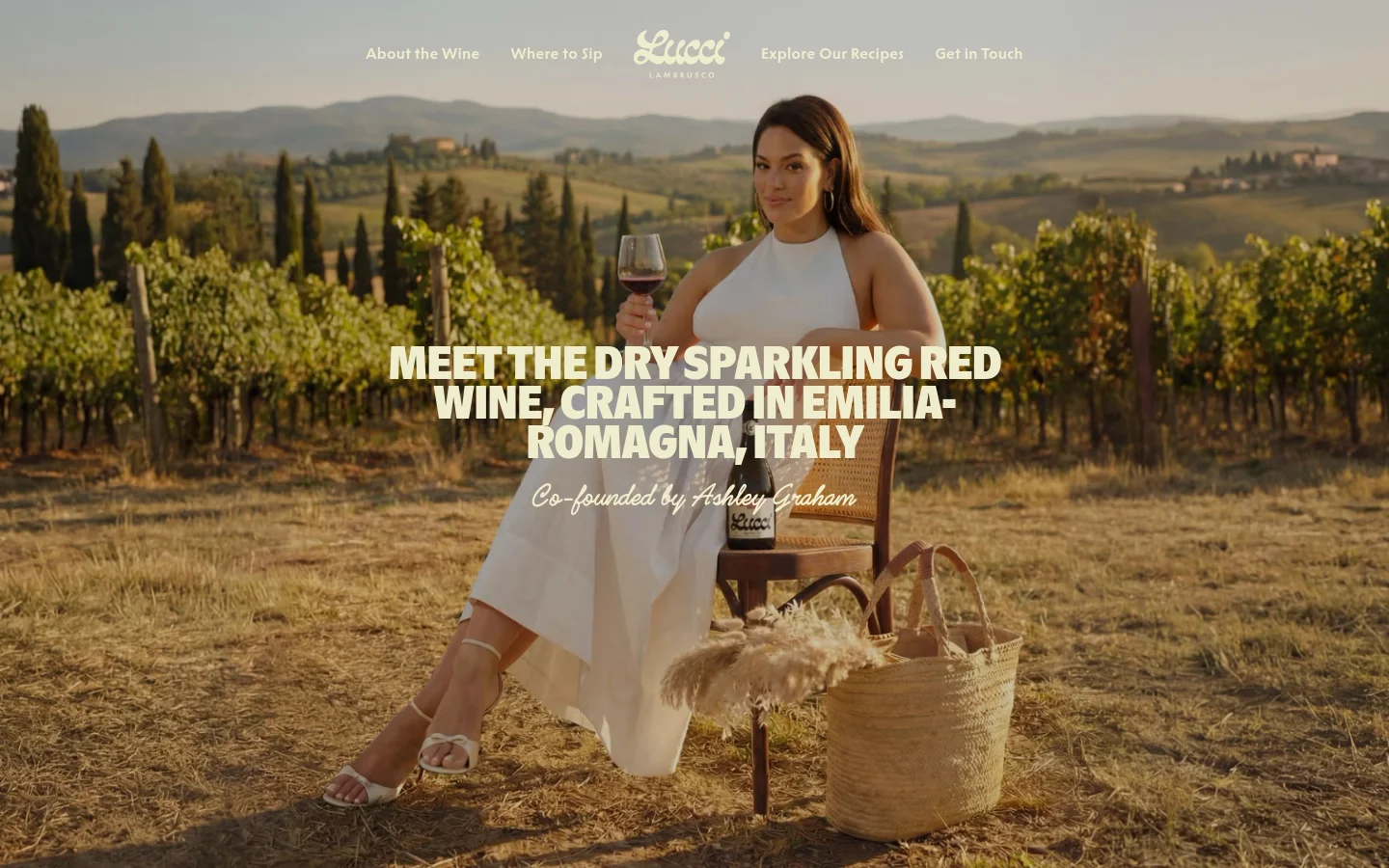 The desktop homepage features a large hero image of Ashley Graham sitting in a vineyard holding a glass of red wine. A navigation menu is at the top with links for About the Wine, Where to Sip, Explore Our Recipes, and Get in Touch. The main headline reads 'MEET THE DRY SPARKLING RED WINE, CRAFTED IN EMILIA-ROMAGNA, ITALY' with a sub-headline mentioning co-founder Ashley Graham.