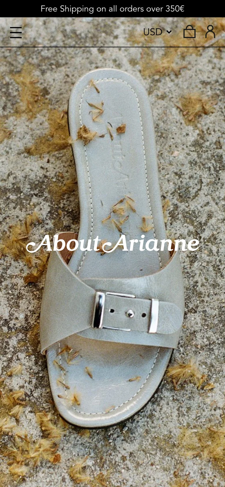 The mobile version presents a vertically oriented hero image focusing closely on a single light grey sandal resting on a textured, petal-strewn ground. The brand name 'About Arianne' is centered over the image, and the navigation is condensed into a hamburger menu icon at the top left, with currency and account icons at the top right.