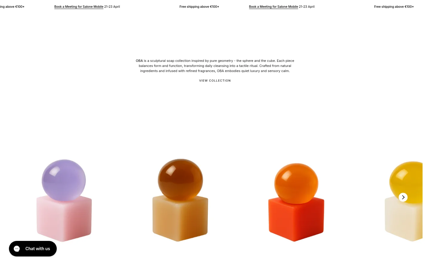 The desktop homepage features a minimalist white layout with a top announcement bar and a centered hero section. The text introduces the 'OBA' sculptural soap series, followed by a 'VIEW COLLECTION' call to action and a row of colorful, geometric soap sculptures in shades of purple, amber, and orange. A 'Chat with us' button is anchored in the bottom left corner.
